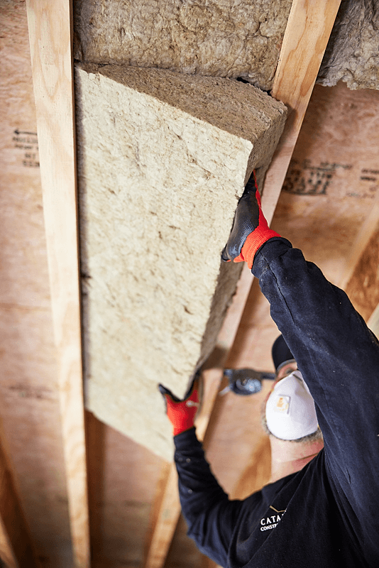 A worker wearing a white face mask, black long-sleeve shirt, and red gloves installs a large rectangular insulation board between wooden framing studs in an attic or wall structure.