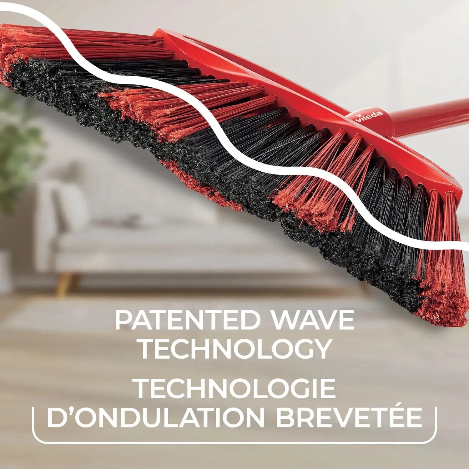 A close-up of the VILEDA 48" Super Angle Broom showcases red and black bristles with “Patented Wave Technology” (English/French), plus a sturdy steel handle, set against a blurred living room background.