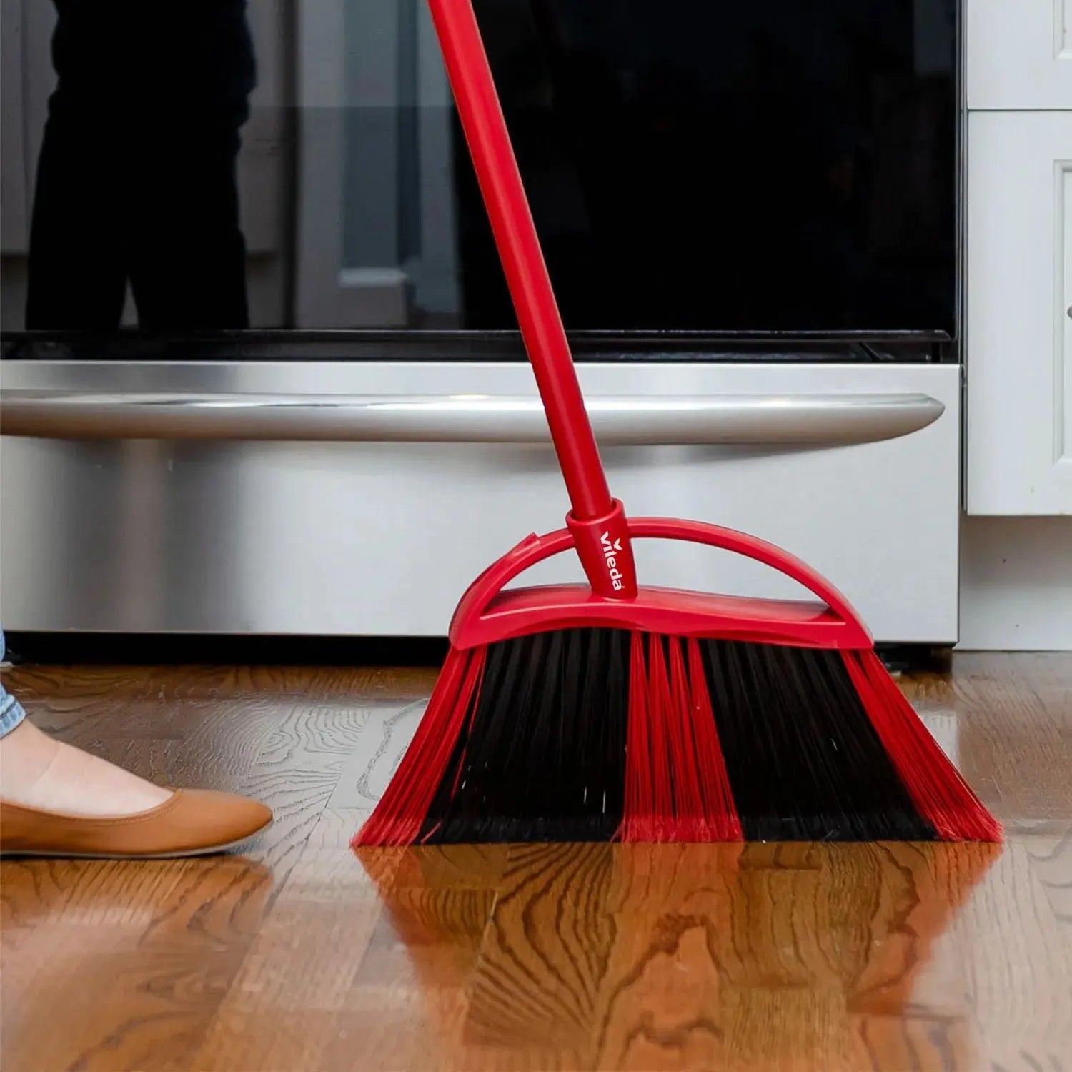 A person in brown flats sweeps a wooden floor with a 48" VILEDA Super Angle Broom, which has plastic bristles and a steel handle, in front of a stainless steel oven.