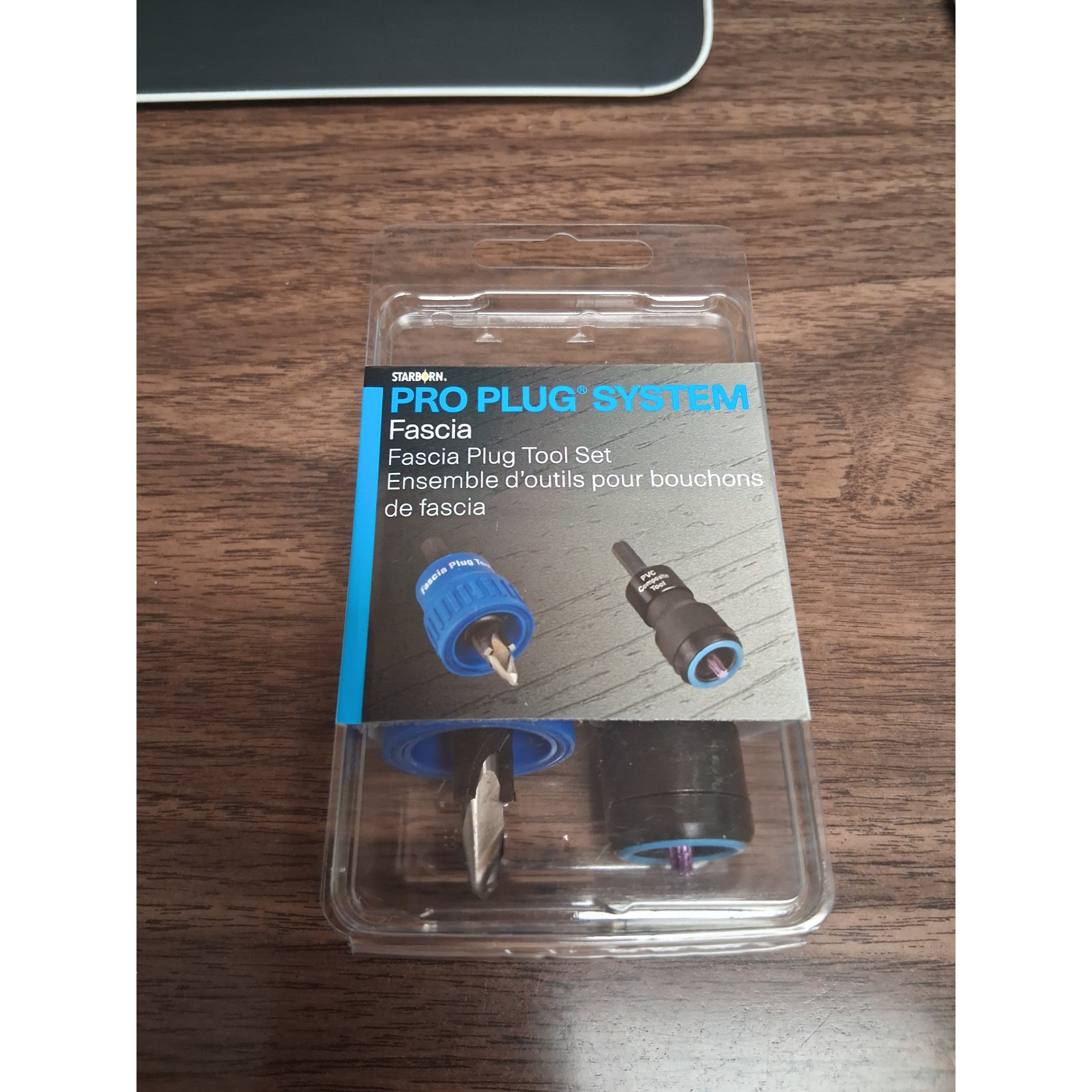 A packaged Trex Starborn Pro-Plug Fascia Tool rests on a wooden desk. Through its clear packaging, two fascia installation tools are visible alongside product details in English and French—making it perfect for efficient fascia board installation.