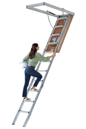A woman in a green sweater climbs the Louisville Ladder Corp Aluminum Attic Ladder-375LB Load Capacity, holding a white bucket in one hand. The 22.5 x 54-inch ladder extends to an open attic hatch in the ceiling above her.