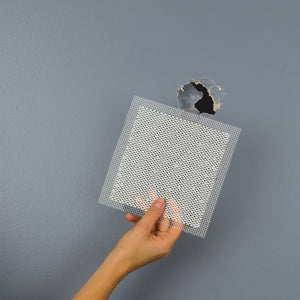 A hand holds a Saint-Gobain Adfors FIBATAPE® 4X4 Aluminum Wall Repair Patch (24 Pack) in front of a hole in a blue wall, ready to cover and repair the damaged area.