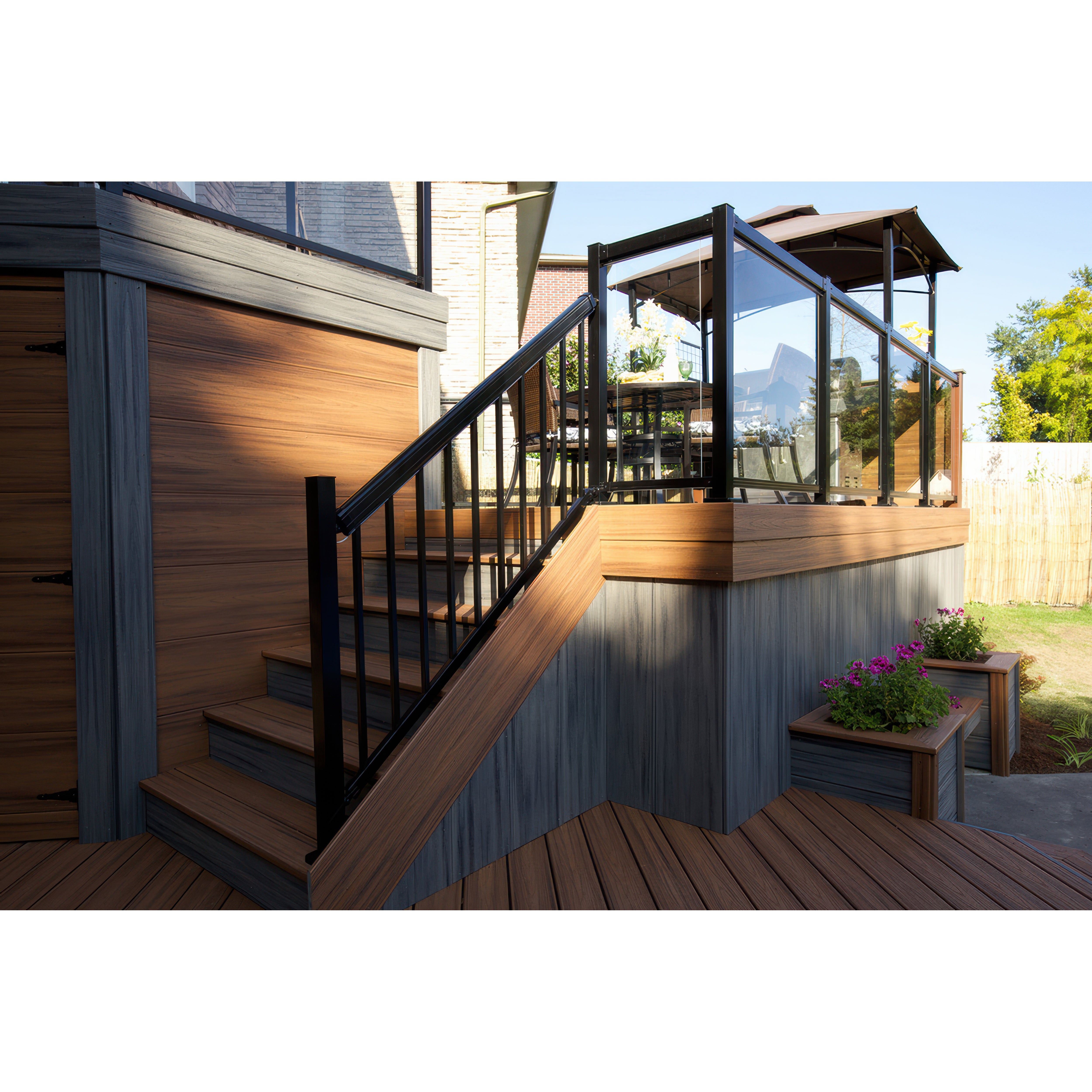 A modern outdoor deck with wood and gray paneling features stairs with sleek aluminum railing, a Regal Rail 36-7/8"x27" clear tempered glass panel enclosure, potted pink flowers, and an inviting seating area under a canopy.