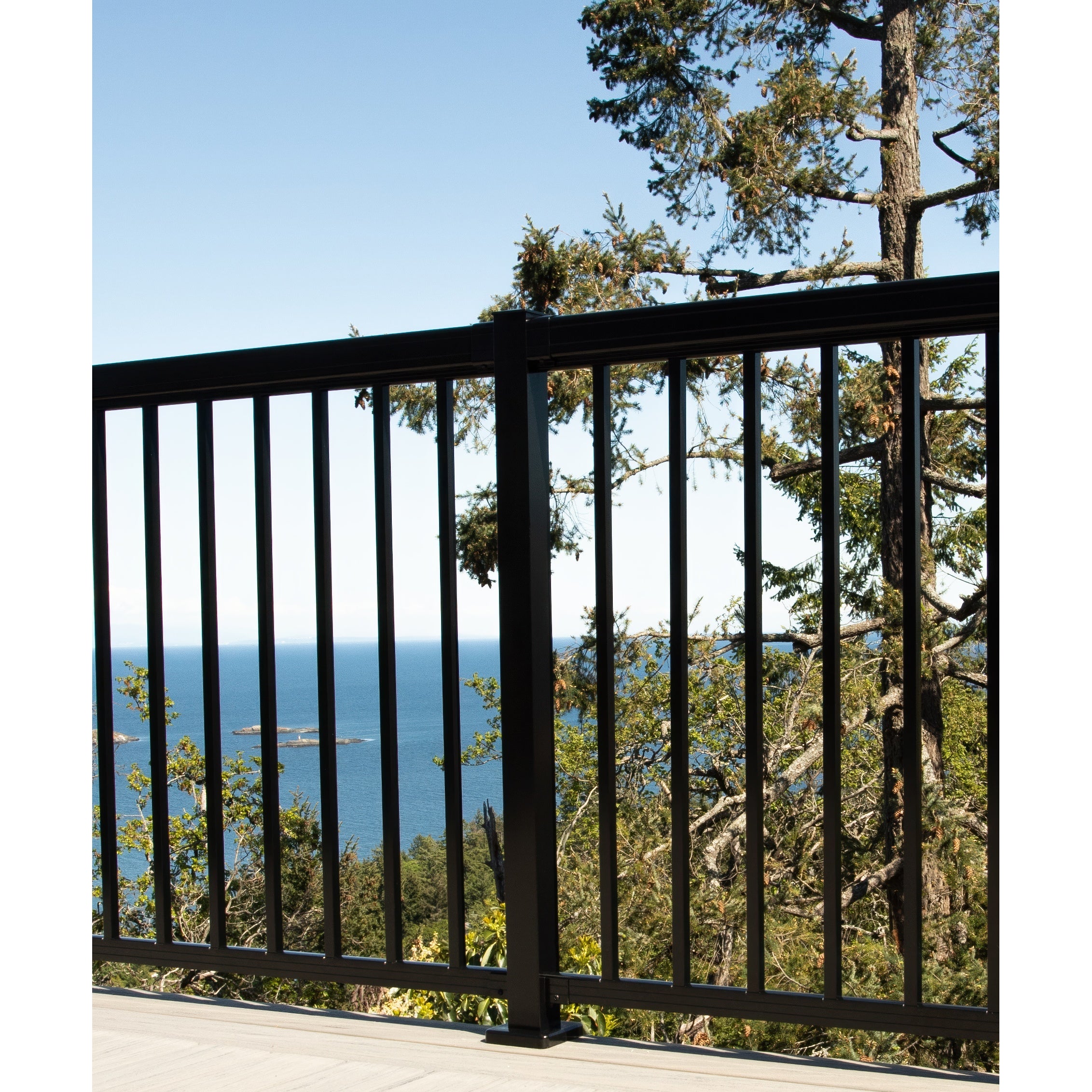 A Regal Rail 6' Straight Pickets railing in textured black by Regal Rail lines the balcony, overlooking trees and a scenic ocean view under a clear blue sky.