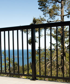 A Regal Rail 6' Straight Pickets railing in textured black by Regal Rail lines the balcony, overlooking trees and a scenic ocean view under a clear blue sky.