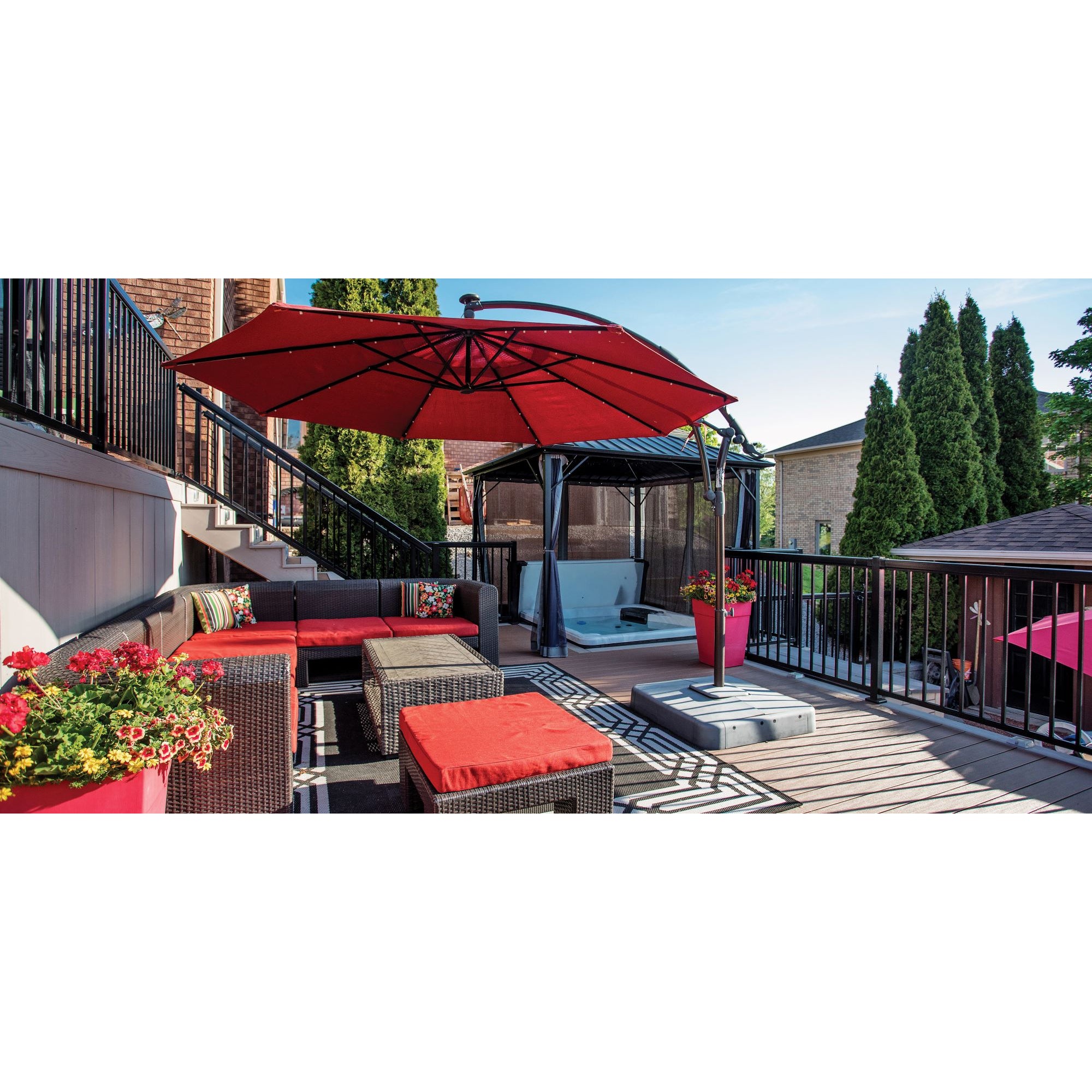 A cozy patio with a red umbrella, wicker sofas with red cushions, potted flowers, and a hot tub under a gazebo, surrounded by Regal Rail 6' Straight Pickets in Textured Black (FOR 42") from Regal Rail and tall green trees.