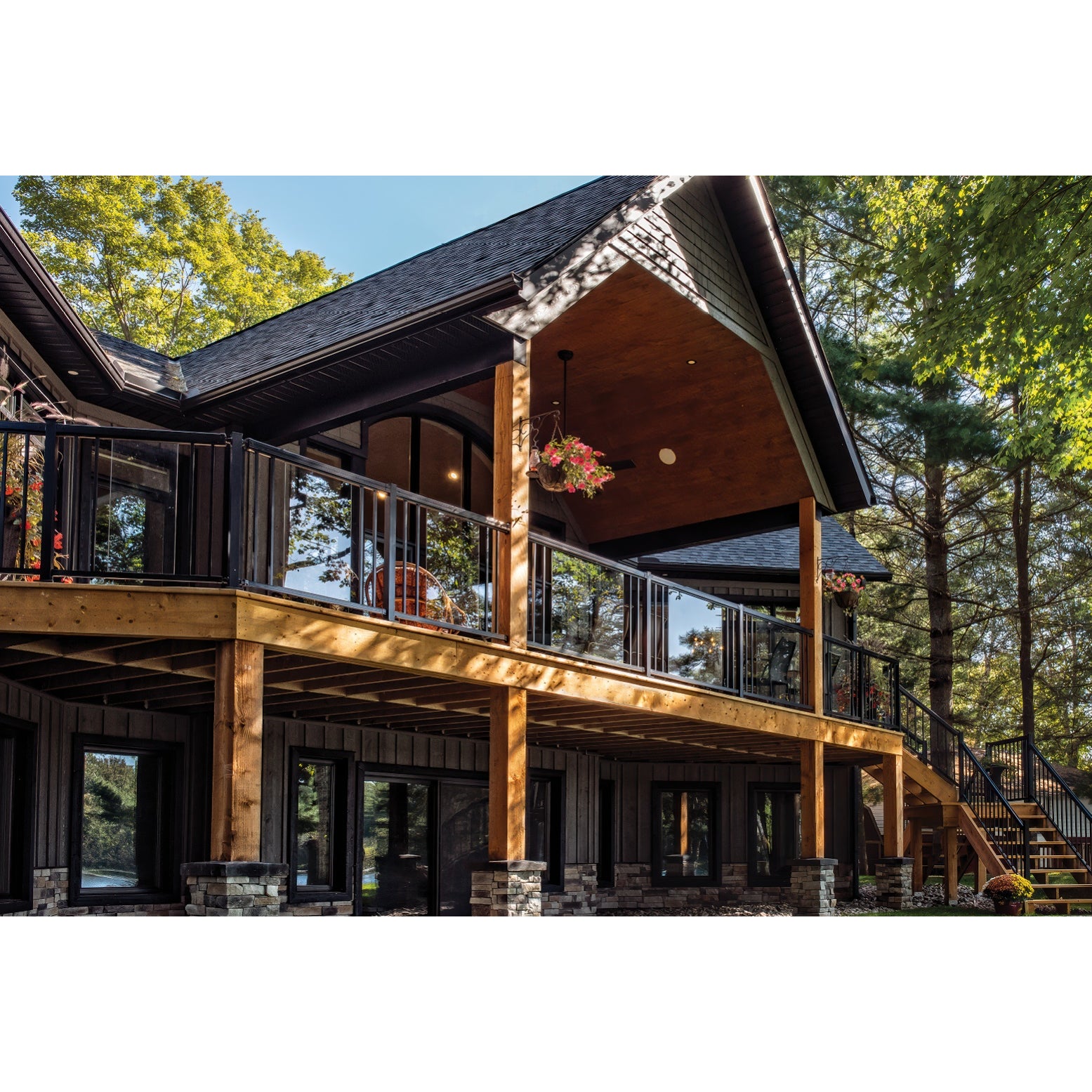 A modern two-story cabin with dark wood siding, large windows, an upper deck featuring Regal Rail 6' Straight Pickets in Textured Black (FOR 42"), hanging flower baskets, a wooden staircase, and surrounded by tall green trees.