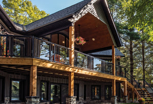 A modern two-story cabin with dark wood siding, large windows, an upper deck featuring Regal Rail 6' Straight Pickets in Textured Black (FOR 42"), hanging flower baskets, a wooden staircase, and surrounded by tall green trees.
