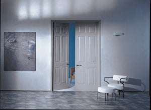 A modern room features partly open Jeld-Wen Colonist 6-Panel Textured Slab double doors (24"x96"x1-3/8"), revealing blue walls, a white armchair with black arms, matching stool, tiled floor, and abstract wall art.