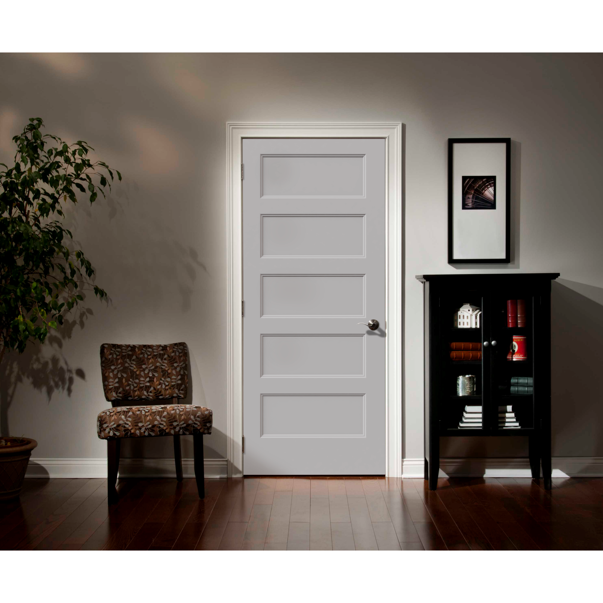 A Jeld-Wen 12"X80"X1-3/8" Conmore 5-Panel Recessed Slab door is centered in the room, with a patterned chair left, tall black cabinet with books and decor right, a plant in the corner, and a framed photo above the cabinet.