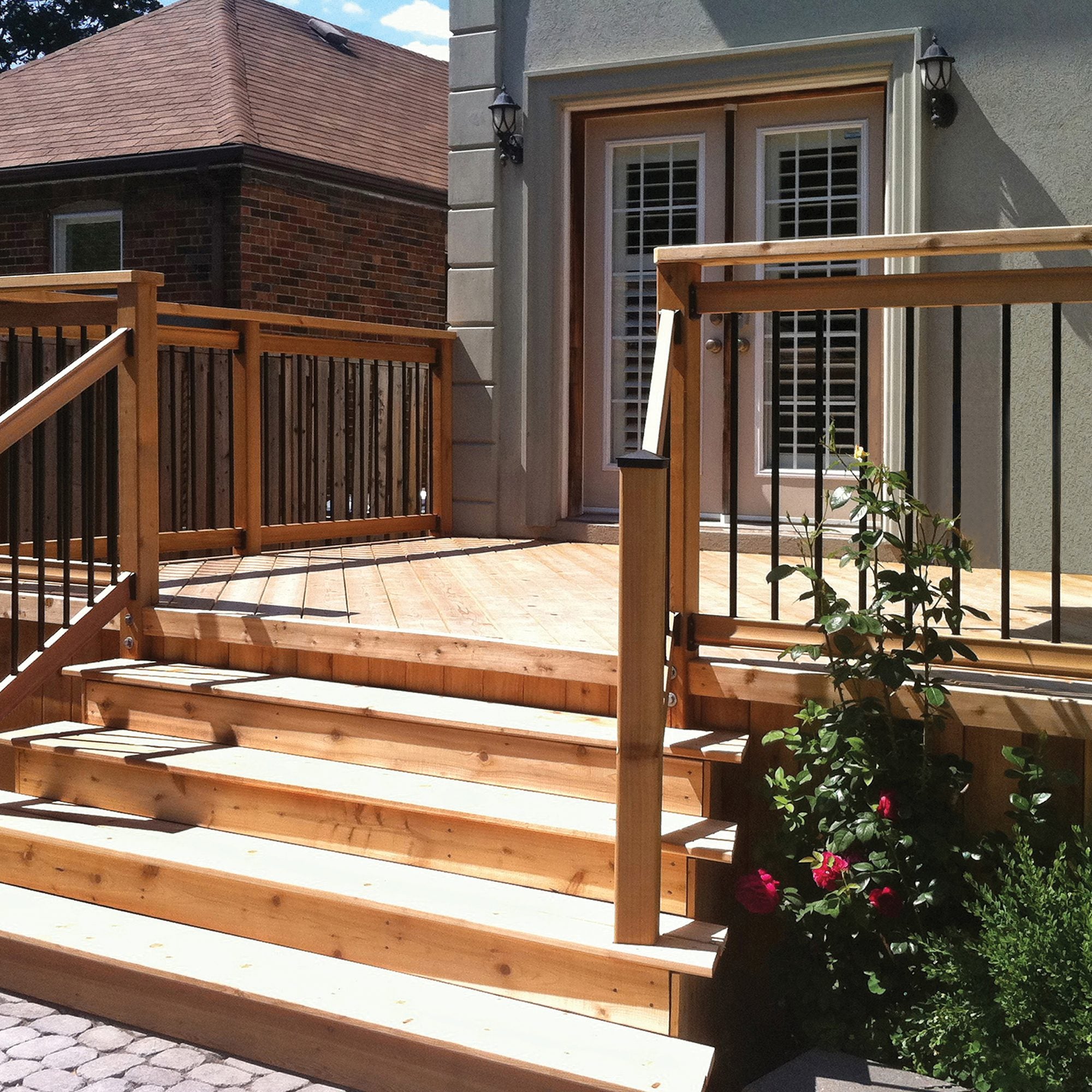 A wooden deck with stairs features Nuvo Iron 3/4"x26" Round Galvanized Steel Balusters in black. The railing leads to glass double doors, red roses grow by the steps, and another building is seen in the background.