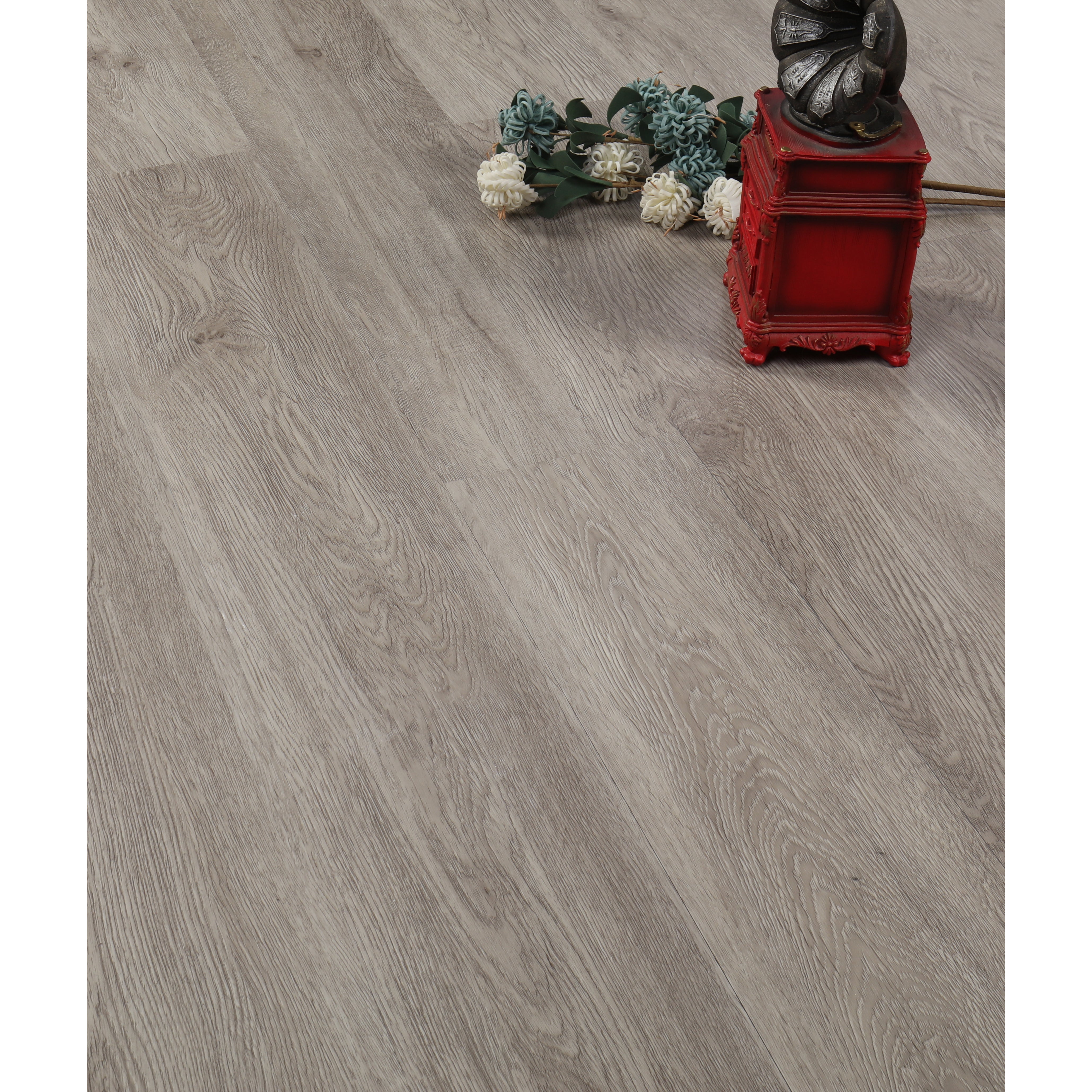 EasyStreet LVP 5MM Flooring, Catherine by Taiga Building Products Ltd features a light gray wood look, shown here with white and blue artificial flowers and a red box with a metallic ornament arranged on the floor.