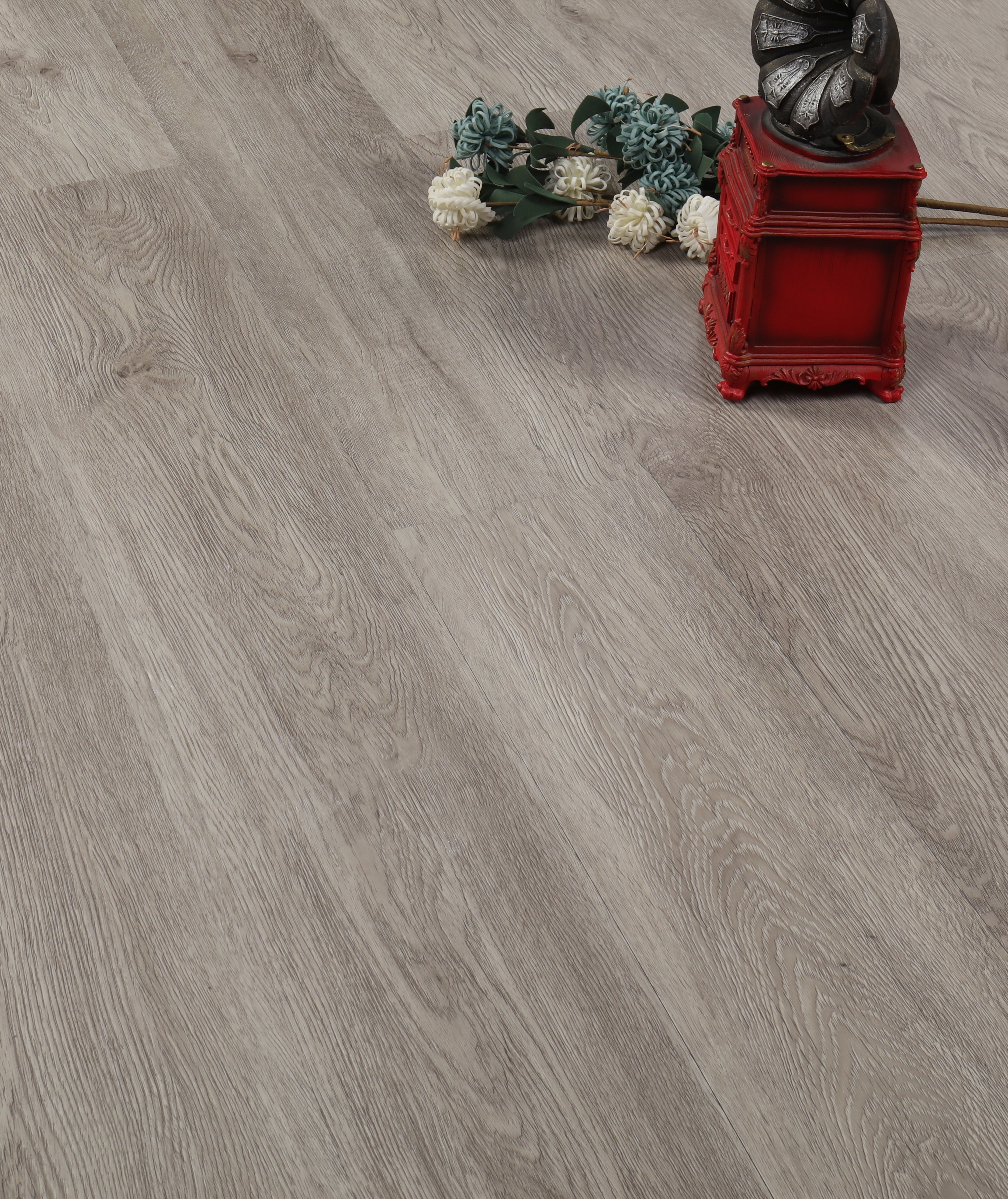 EasyStreet LVP 5MM Flooring, Catherine by Taiga Building Products Ltd features a light gray wood look, shown here with white and blue artificial flowers and a red box with a metallic ornament arranged on the floor.