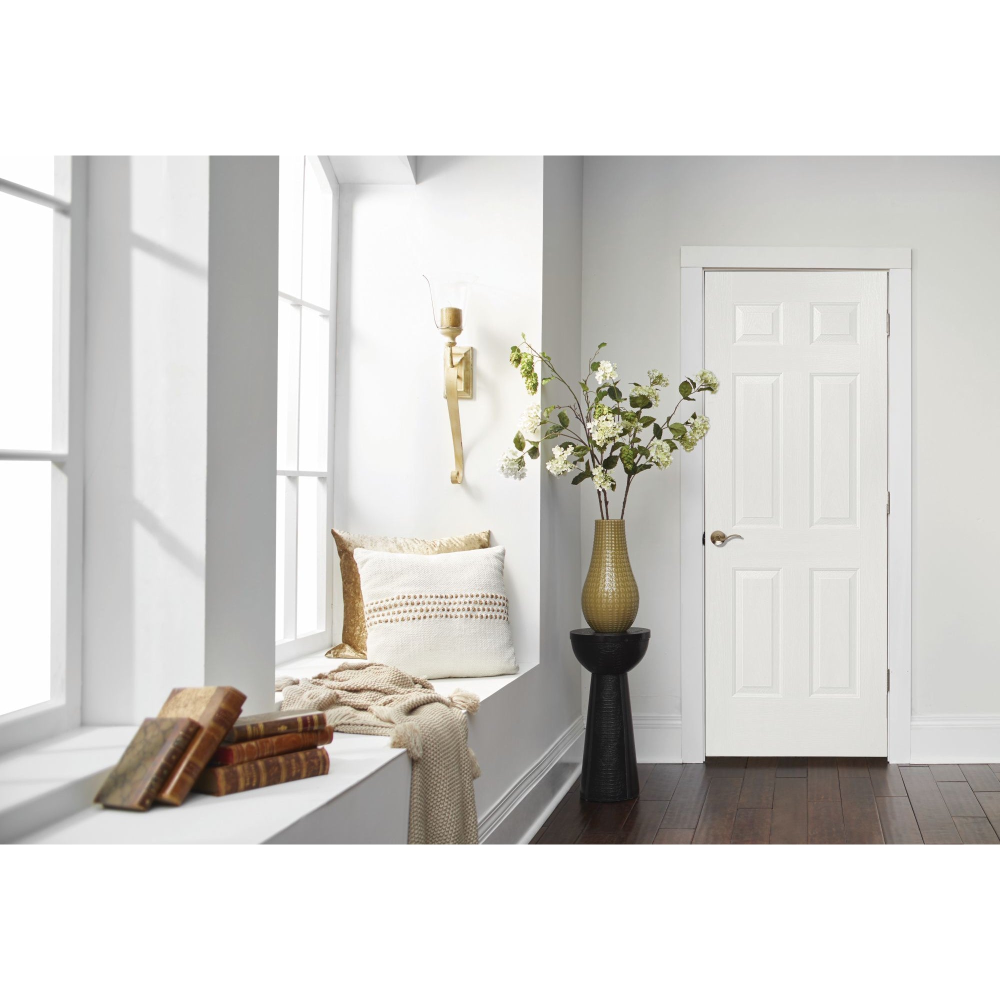 A bright hallway features large windows, a cushioned window seat, beige throw, stacked books, a modern sconce, tall vase with flowering branches beside a Jeld-Wen Colonist 26"X96" 6-panel textured slab interior door.