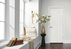 A bright hallway features large windows, a cushioned window seat, beige throw, stacked books, a modern sconce, tall vase with flowering branches beside a Jeld-Wen Colonist 26"X96" 6-panel textured slab interior door.