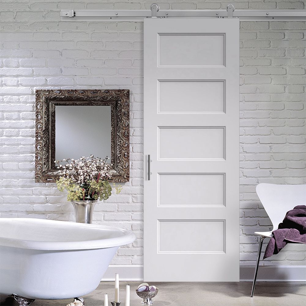 A bathroom featuring a Jeld-Wen 12"X80"X1-3/8" Conmore 5-Panel Recessed Slab door on a metal track, white freestanding tub, floral-accented mirror, white brick walls, and a modern chair with a purple towel.