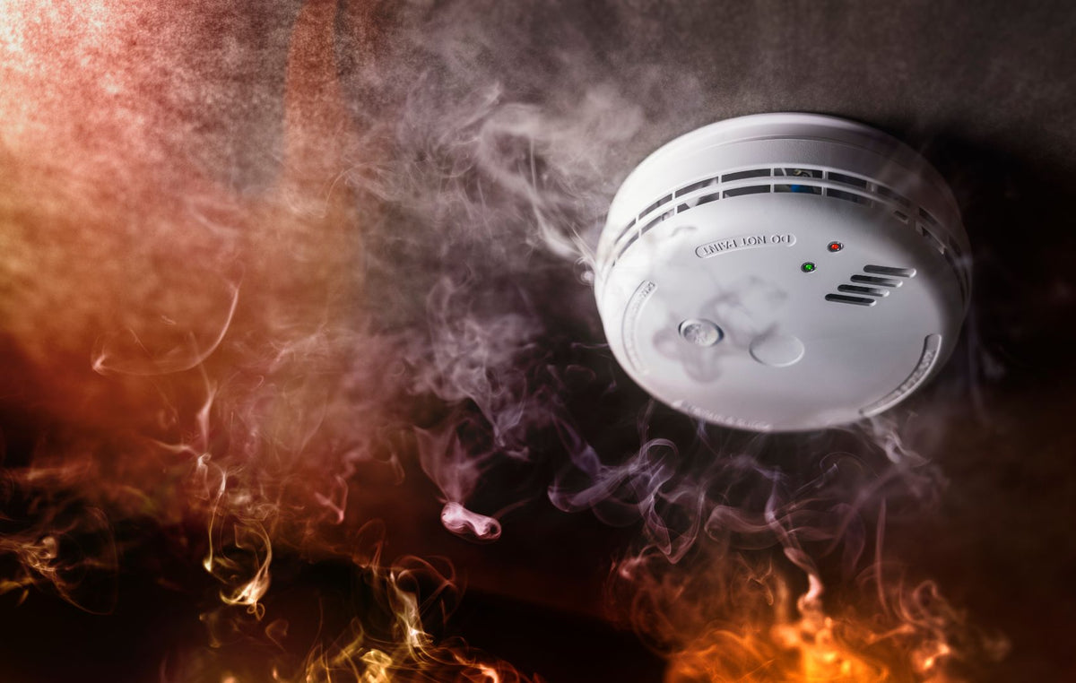 A white round smoke detector shown against a dark background with dramatic orange flames and smoke effects.