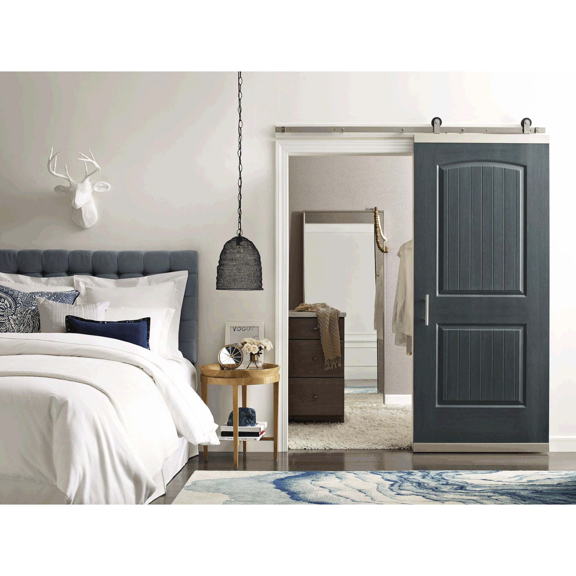 A modern bedroom features a blue upholstered bed, white bedding, round wood side table, black woven hanging lamp, and a Jeld-Wen 12"x80"x1-3/8" Santa Fe 2-Panel Slab barn door partially open to reveal a dresser and mirror.