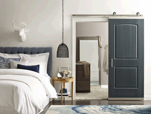 A modern bedroom features a blue upholstered bed, white bedding, round wood side table, black woven hanging lamp, and a Jeld-Wen 12"x80"x1-3/8" Santa Fe 2-Panel Slab barn door partially open to reveal a dresser and mirror.