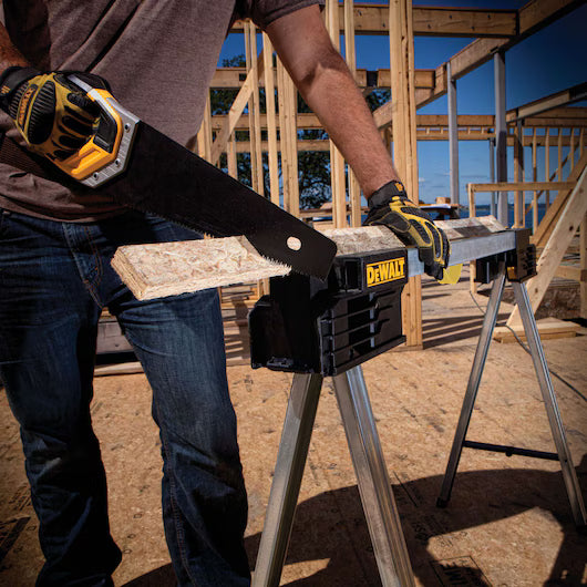 A person wearing gloves uses a hand saw to cut a wooden board on a DEWALT Dewalt Metal Folding Sawhorse at a construction site with wooden framing in the background.