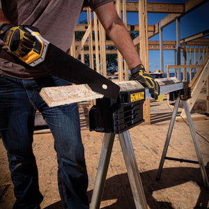 A person wearing gloves uses a hand saw to cut a wooden board on a DEWALT Dewalt Metal Folding Sawhorse at a construction site with wooden framing in the background.