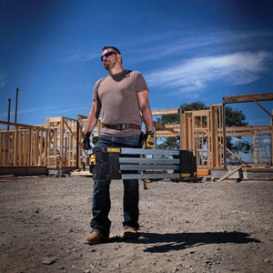 A man in work gloves and sunglasses holds a DEWALT Metal Folding Sawhorse at a construction site with wooden framing and a clear blue sky in the background.
