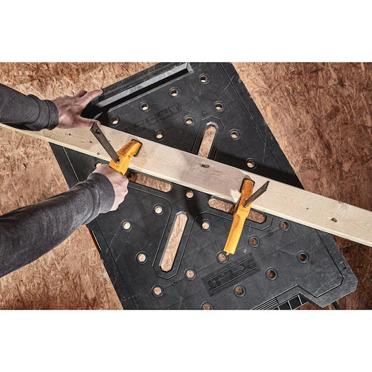 A person secures a wooden board to a Dewalt Folding Workbench from TurkstraLumberLtd using two yellow clamps, getting ready for a woodworking project.
