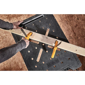 A person secures a wooden board to a Dewalt Folding Workbench from TurkstraLumberLtd using two yellow clamps, getting ready for a woodworking project.