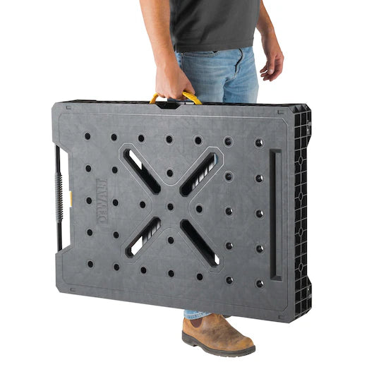 A person carrying the Dewalt Folding Workbench by TurkstraLumberLtd—a black, rectangular portable workbench featuring a yellow handle, multiple surface holes, and X-shaped grooves.