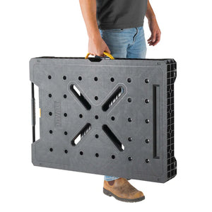 A person carrying the Dewalt Folding Workbench by TurkstraLumberLtd—a black, rectangular portable workbench featuring a yellow handle, multiple surface holes, and X-shaped grooves.