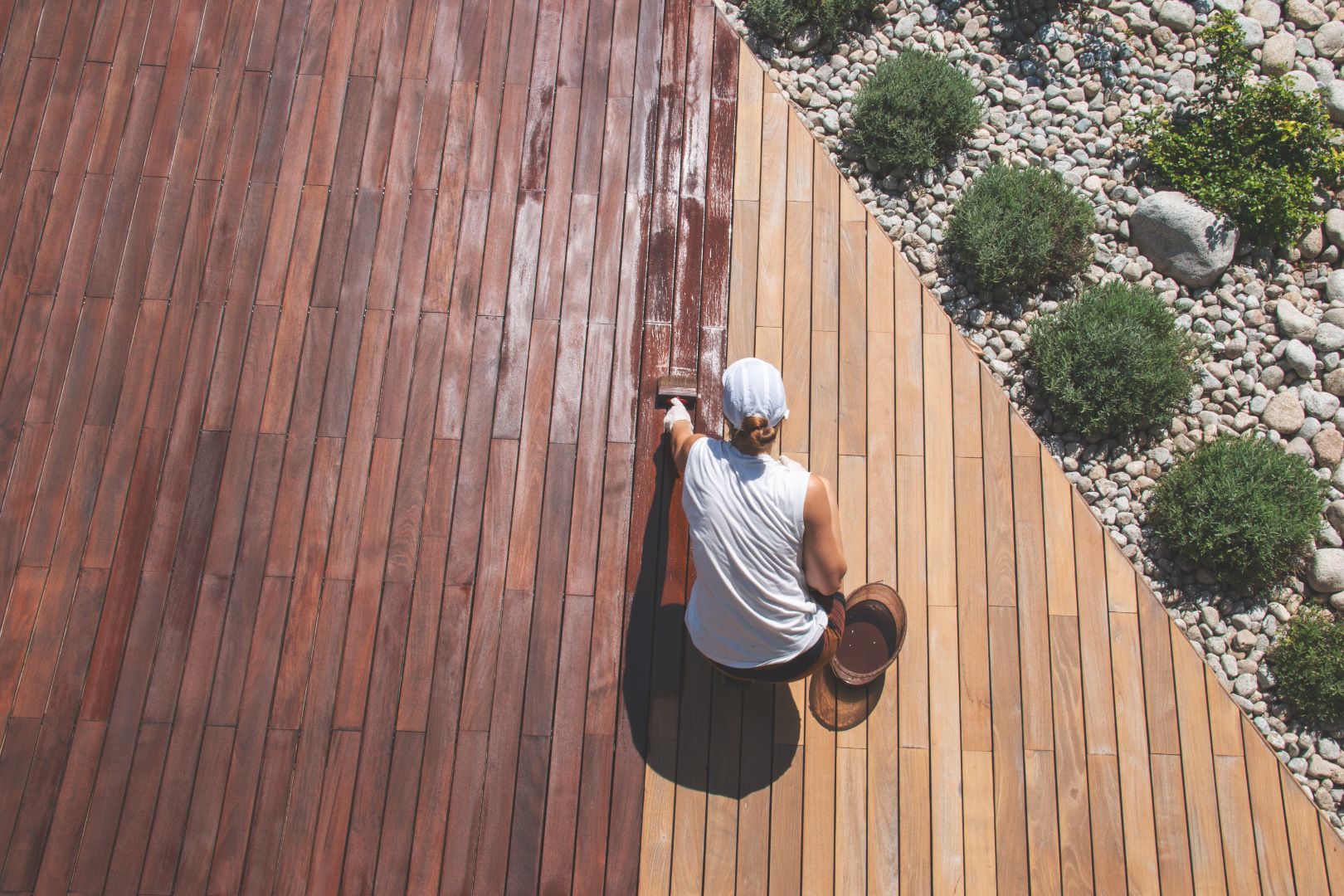 A woman staining her deck a dark brown among a shrub and rock garden