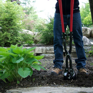 Person using hole digging tool in a garden setting with plants and rocks in the background