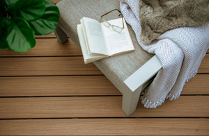 A cozy setup with an open book, glasses, and a striped blanket on a cushioned bench sits by a green plant atop Trex 1" x 6" x 20' Enhance Naturals Grooved deck boards in Golden Hour from Trex.