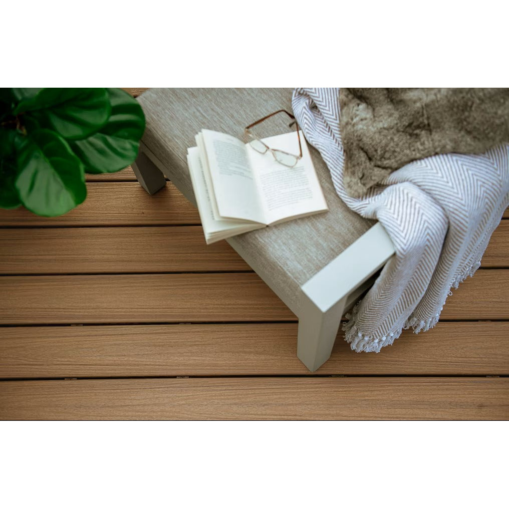 An open book with glasses sits on a cushioned bench beside a striped blanket and green plant, all set on Trex Enhance Naturals Golden Hour 1" x 12" x 12' composite deck boards by Trex for easy, low-maintenance outdoor living.