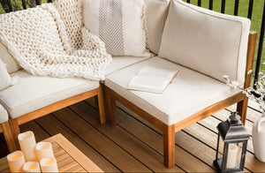 A cozy outdoor seating area with cream cushions and a black lantern sits atop 1" x 6" x 20' Trex Enhance Naturals Grooved decking in Golden Hour by Trex, with a green lawn visible beyond the railing.