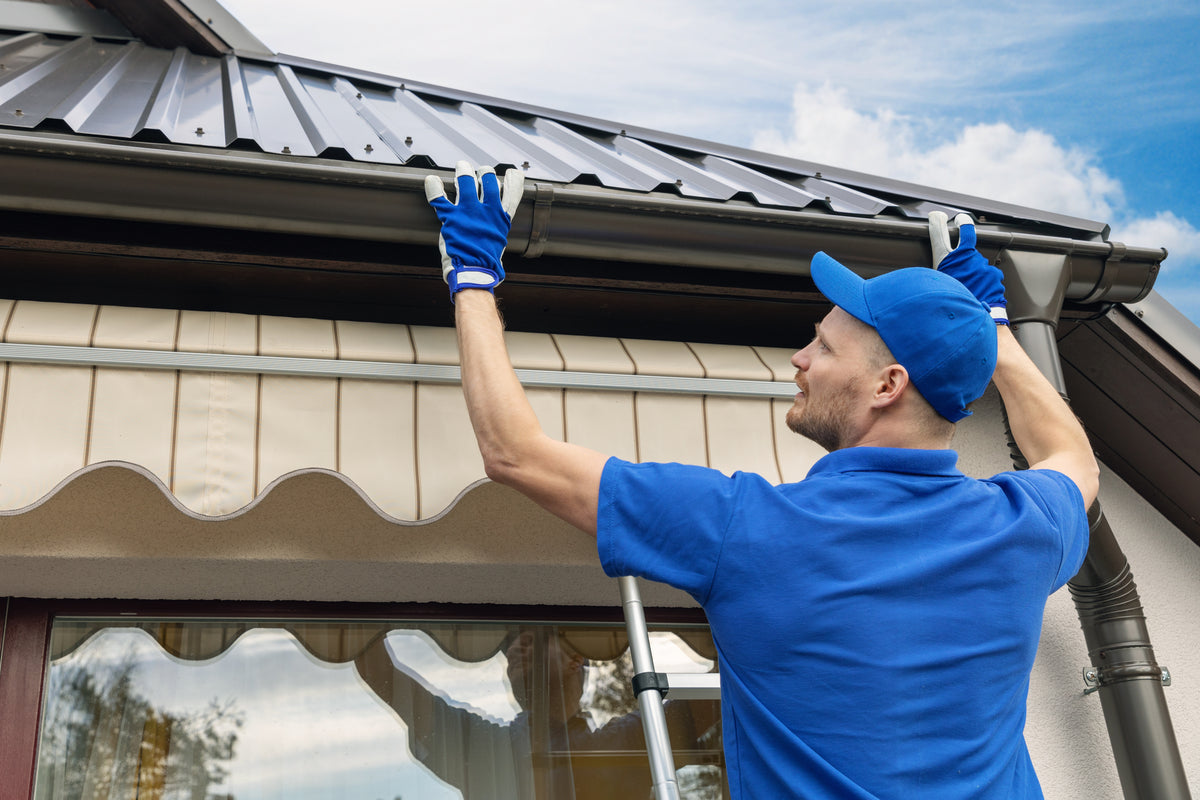 A man in a blue shirt and cap wearing blue work gloves adjusts a gutter downspout on a house roof under a dark metal tile, with a beige awning and blue sky in the background.