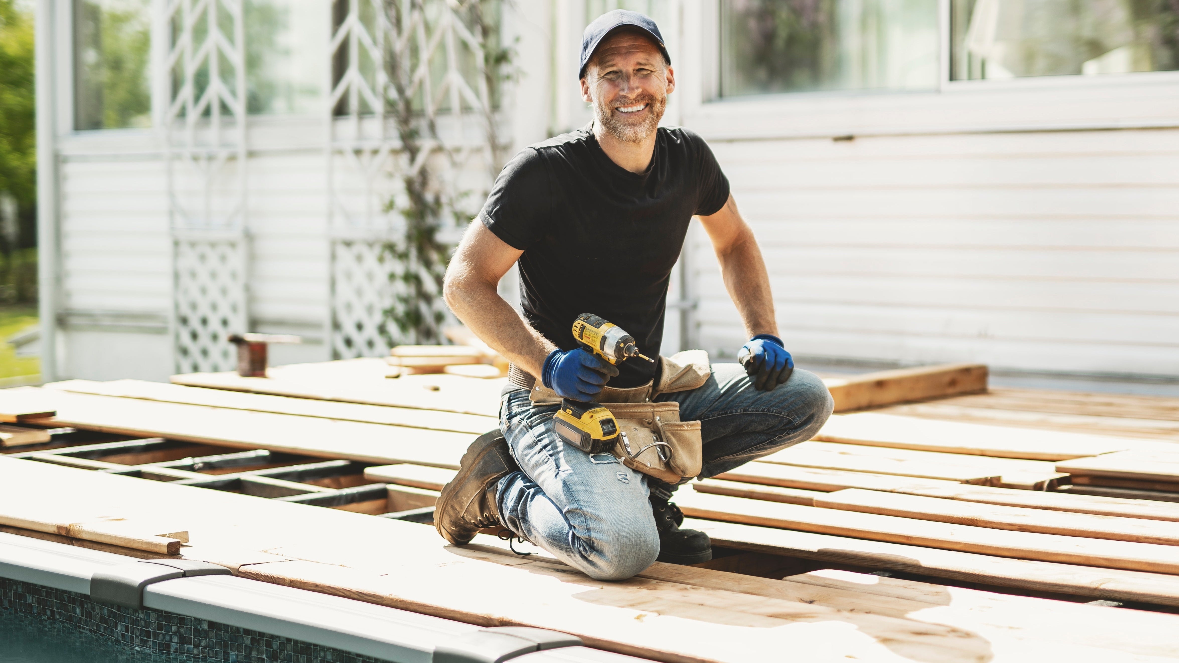 Man working on a wooden deck with tools, wearing a black t-shirt and blue gloves.