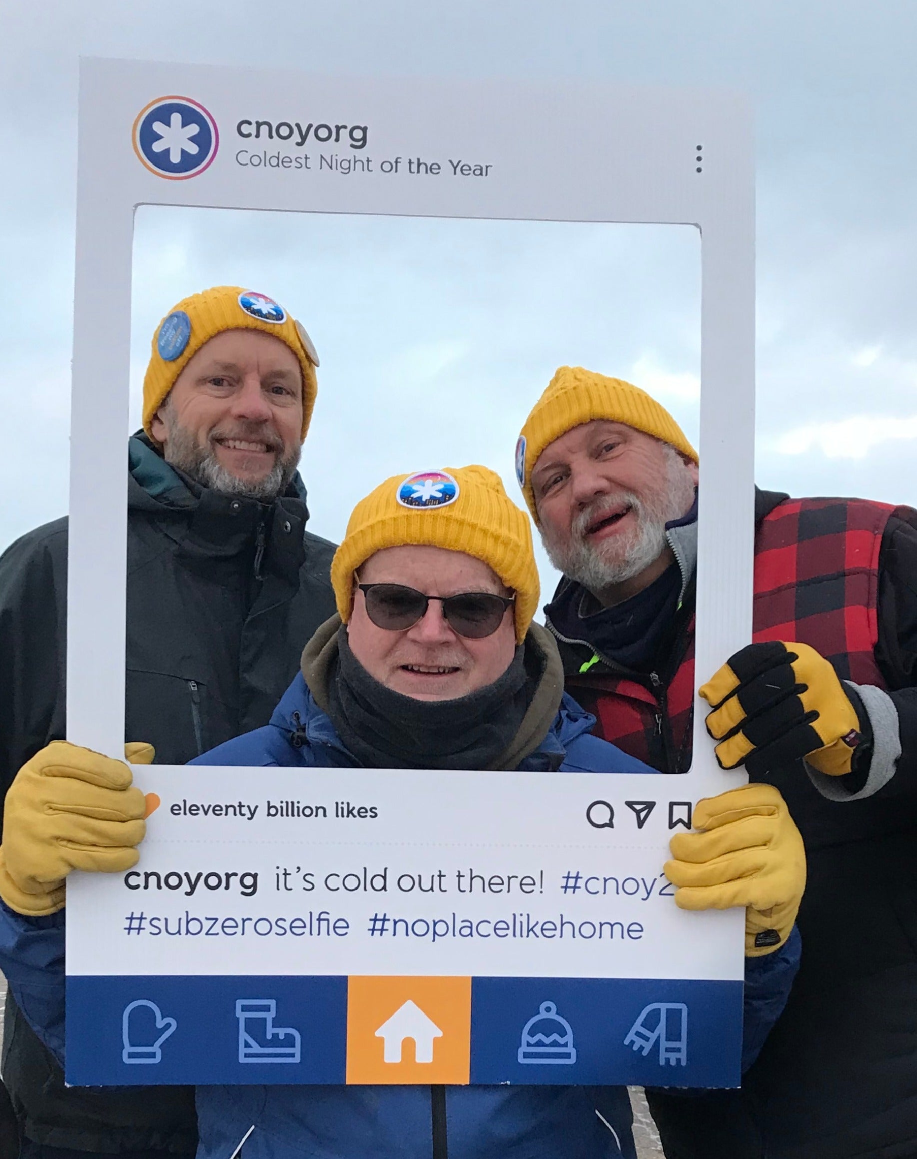 Three smiling people in yellow hats and gloves pose outdoors, holding a large Instagram-style frame promoting Coldest Night of the Year with hashtags and the CNOY logo. The sky is overcast.