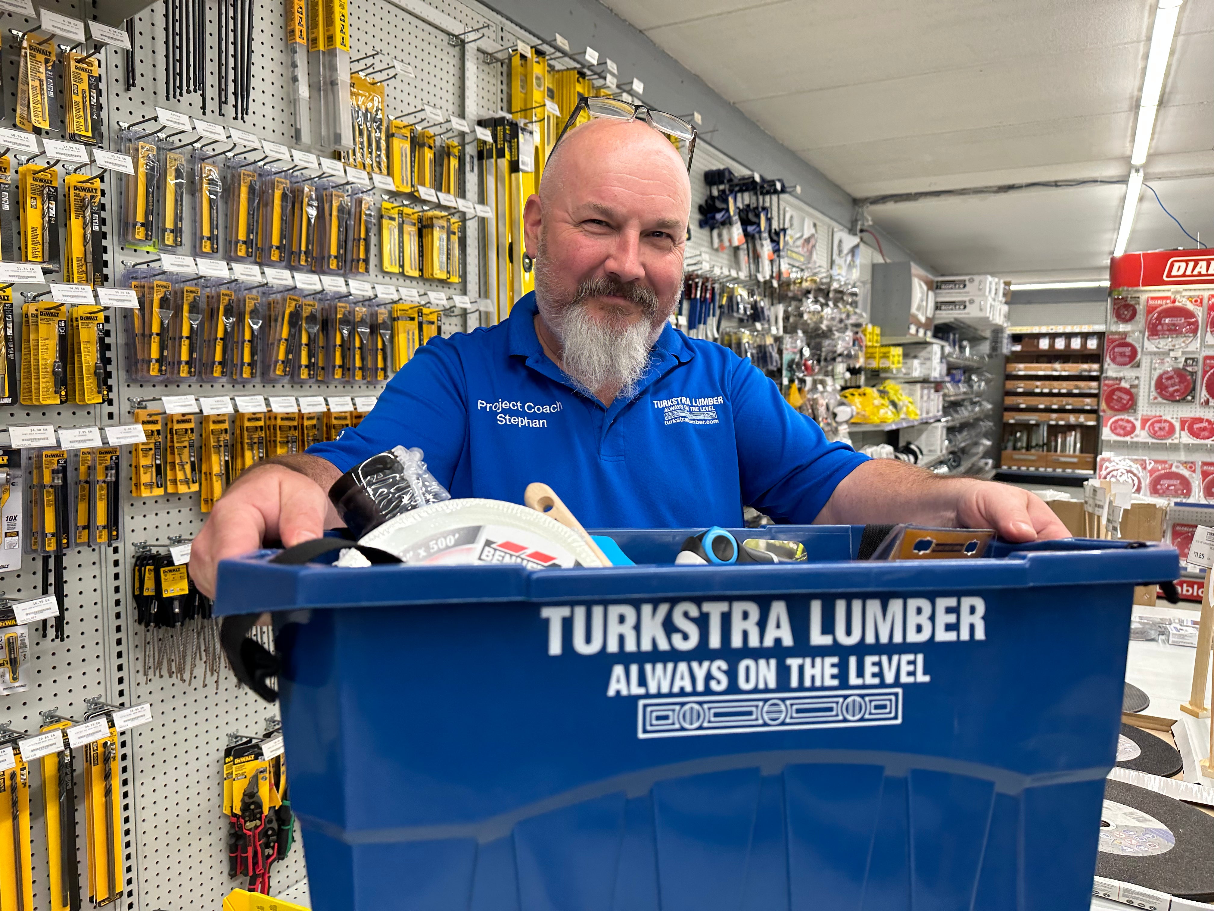 A man in a blue Turkstra Lumber shirt stands in a hardware store aisle, smiling and holding a blue bin filled with tools and supplies. Shelves with various tools hang on the wall behind him.