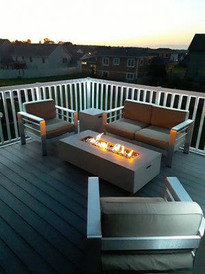 Modern patio furniture surrounds a rectangular fire pit on a deck with Regal Rail 6' Straight Pickets, White (FOR 42") by Regal Rail, at sunset, with softly lit houses and sky in the background.