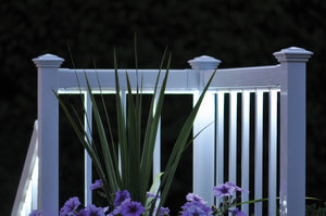 The Regal Rail 6' Straight Pickets in white by Regal Rail are lit up at night, standing out behind green leaves and purple flowers, highlighting their modern elegance against a dark, blurred background.
