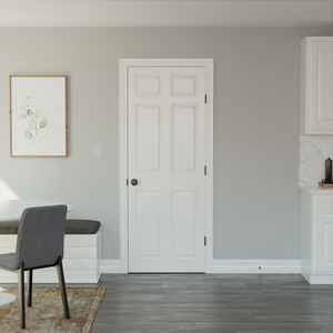 A Jeld-Wen 24"x96"x1-3/8" Colonist 6-panel textured slab door with a black knob is centered on a light gray wall, flanked by a framed botanical print and gray bench to the left, and white kitchen cabinets to the right.