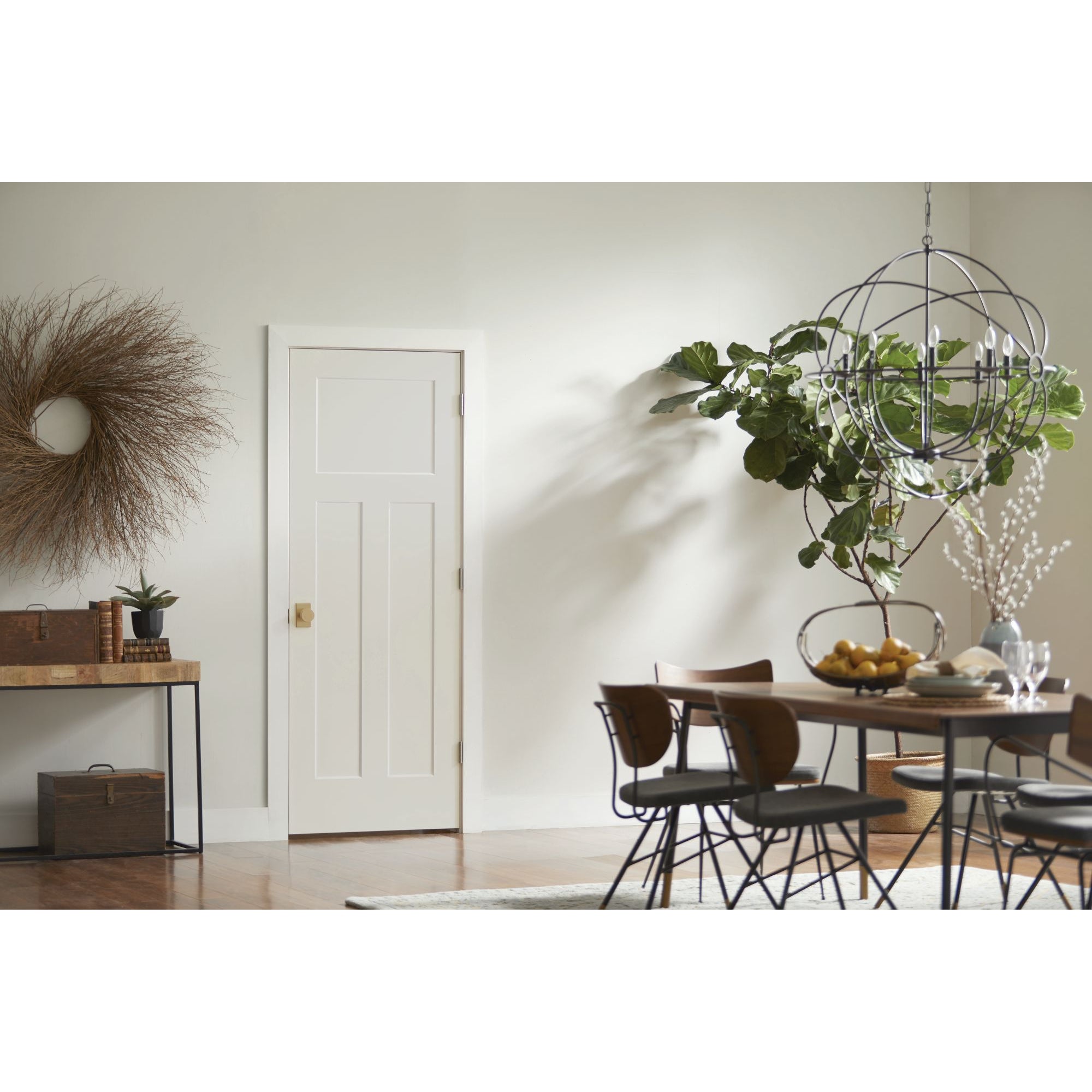 A bright, modern dining area with a large potted plant, round chandelier, wooden table and chairs, console with books and plants, circular wall wreath, and a Jeld-Wen 12"x80"x1-3/8" Craftsman 3-Panel Recessed Slab door.