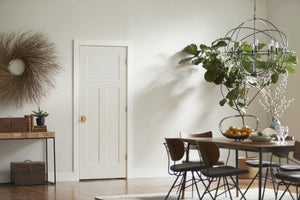 A bright, modern dining area with a large potted plant, round chandelier, wooden table and chairs, console with books and plants, circular wall wreath, and a Jeld-Wen 12"x80"x1-3/8" Craftsman 3-Panel Recessed Slab door.