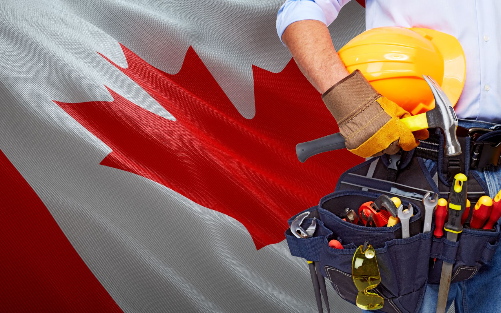 A construction worker fitted with a hard hat, tool belt and lots of tools standing proudly in front of a Canadian flag
