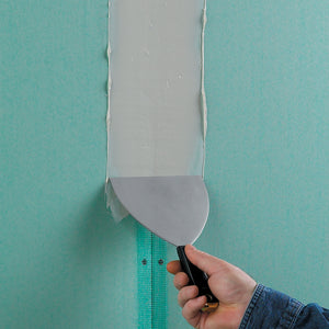 A hand uses a putty knife to smooth joint compound over seams taped with Saint-Gobain ADFORS FibaTape® 1-7/8 x 300' Ultra Thin Mould-X10™ Mold Resistant Drywall Tape on green drywall, enhancing moisture and mold resistance.