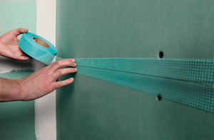 A person applies Saint-Gobain Adfors FIBATAPE® 1-7/8" x 300' Ultra Thin Mould-X10™ Mould Resistant Drywall Tape to the seam of green drywall, preparing it for joint compound and enhancing moisture and mold resistance.