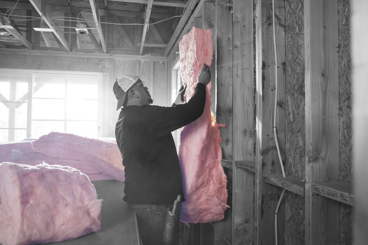 A person in a construction setting hangs a pink insulation panel on a wooden wall frame, with other pink insulation rolls visible in a partially finished room.