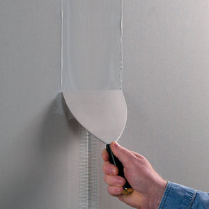 A hand uses a putty knife to apply joint compound over Saint-Gobain Adfors FIBATAPE® 1-7/8" x 300' Ultra Thin Drywall Tape, White, on a wall, smoothing the surface for a clean, seamless finish.