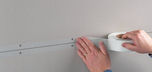 A person applies Saint-Gobain ADFORS FibaTape® 1-7/8" x 300' Ultra Thin Drywall Tape, White, over a joint between two drywall panels, securing it by hand while unrolling the tape from the spool.